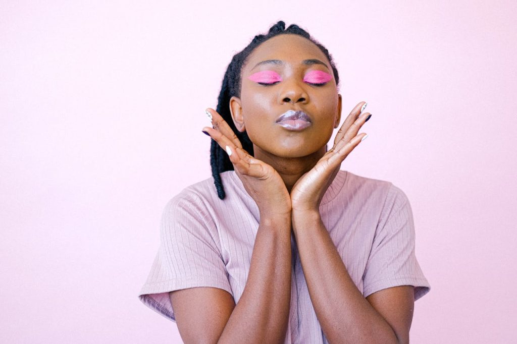 Close-up of a stylish woman posing with closed eyes and pink eye makeup against a pink background.